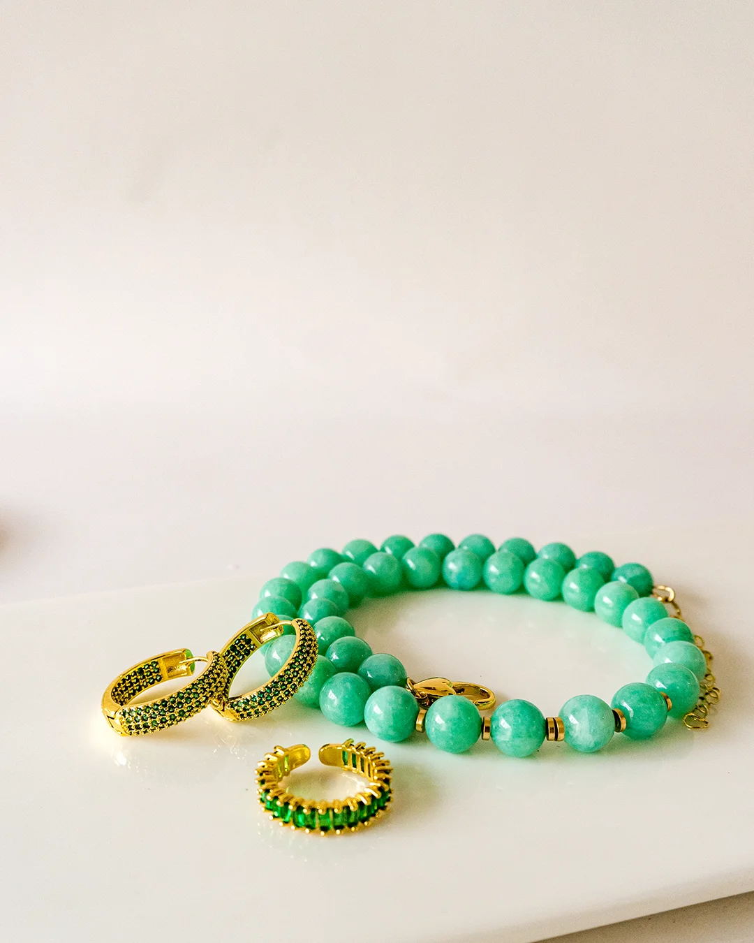 Pulsera verde, anillo dorado y aretes sobre una mesa blanca.